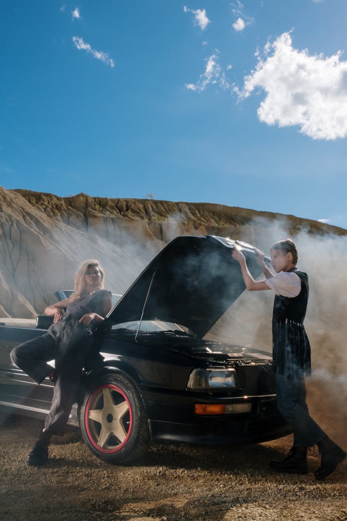 A man and woman by a smoking car with open hood, set in a desert landscape.