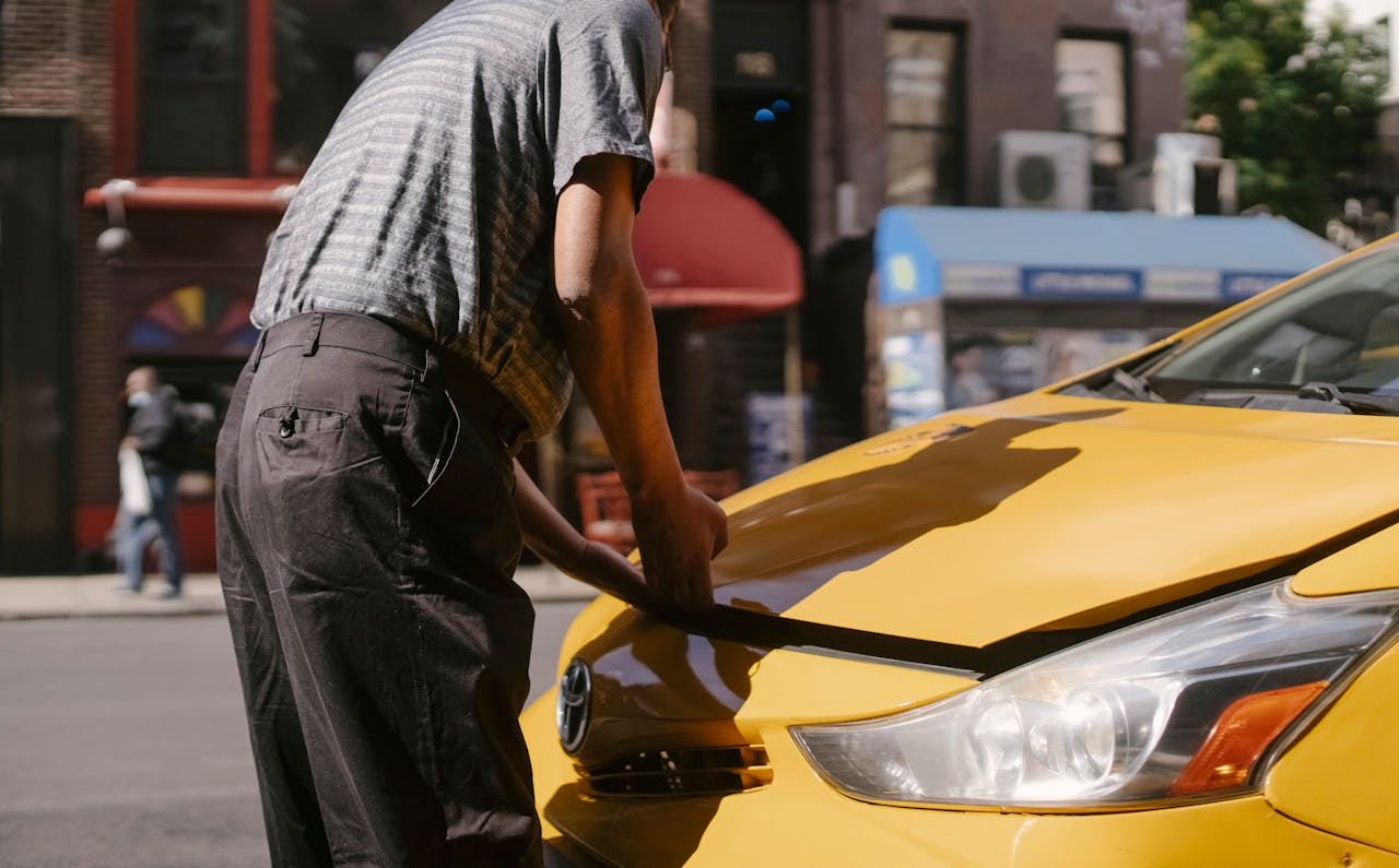 who-we-are Man inspecting open hood of a yellow taxi on city street.