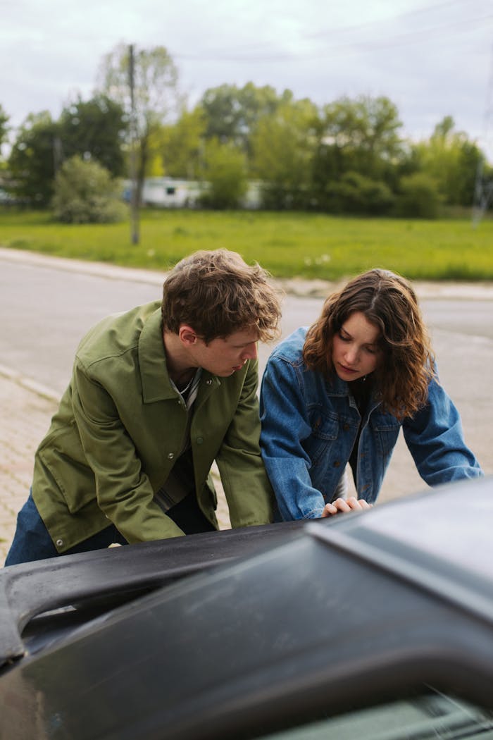 Young couple in denim jackets leaning on a car by a roadside, captured outdoors.